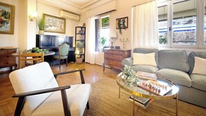 A living room with a sofa, armchair, coffee table, TV, bookshelf, and a window with a view. - Art Gallery Hotel (Athens)