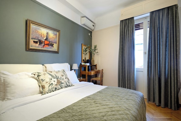 A neatly made bed with a quilt, a desk with a lamp, a painting on the wall, and a window with curtains. - Art Gallery Hotel (Athens)