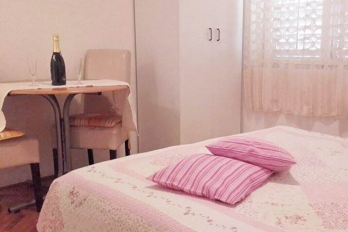 Une pièce comprenant un lit, une chaise, une table avec une bouteille et un verre, et une fenêtre ornée de rideaux en dentelle.