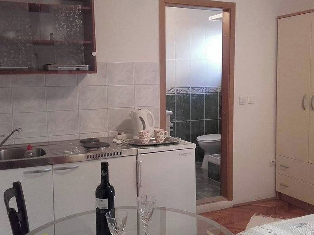 A compact kitchen with a sink, a kettle, a small refrigerator, a dining table with a bottle and glasses, and a view into the adjacent bathroom.