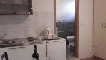 A compact kitchen with a sink, a kettle, a small refrigerator, a dining table with a bottle and glasses, and a view into the adjacent bathroom.