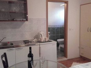 Een compacte keuken met een spoelbak, een waterkoker, een kleine koelkast, een eettafel met een fles en glazen, en uitzicht op de aangrenzende badkamer.