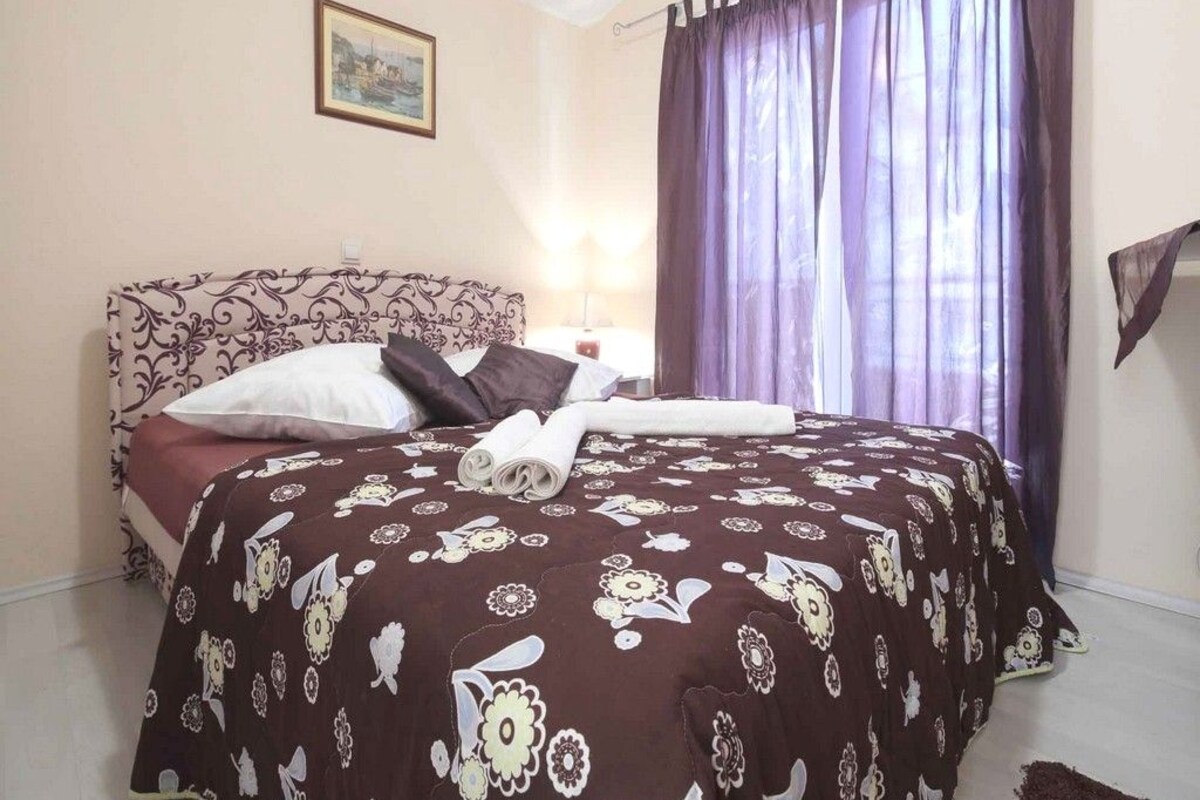 Un dormitorio con una colcha estampada, un cabecero, un cuadro enmarcado en la pared y una ventana con cortinas.