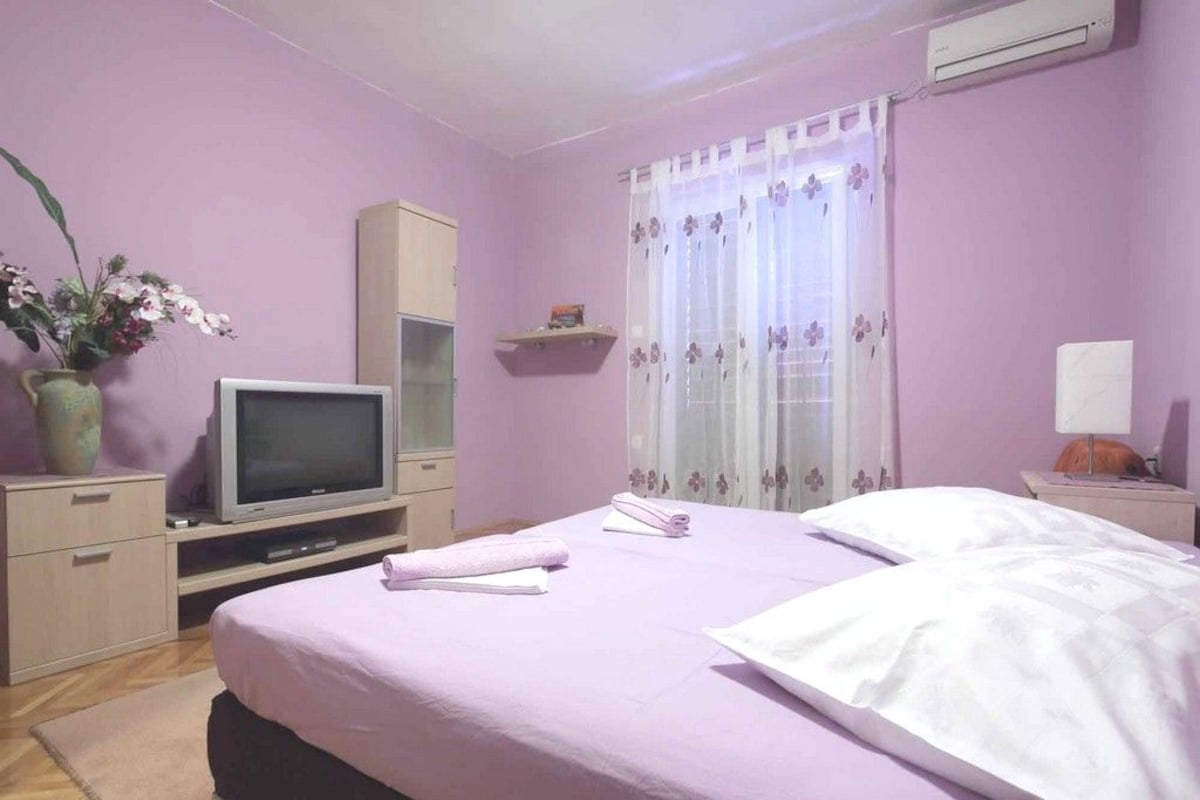 Un dormitorio con cama, televisor, aparador, ventana con cortinas y planta en maceta.