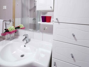 A bathroom with a white sink, a mirror, a shower, and a storage cabinet. - Apartments Goran (Makarska)