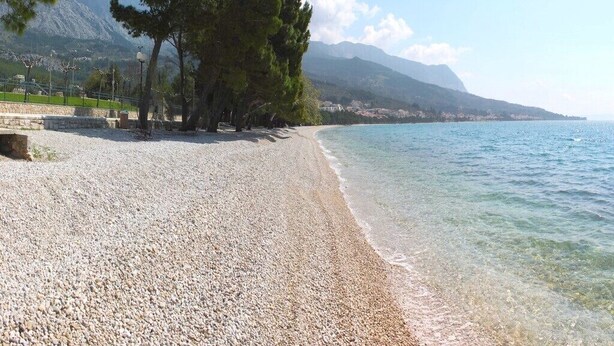 A pebble beach with clear water, trees, and mountains in the background. - Apartments Goran (Makarska)