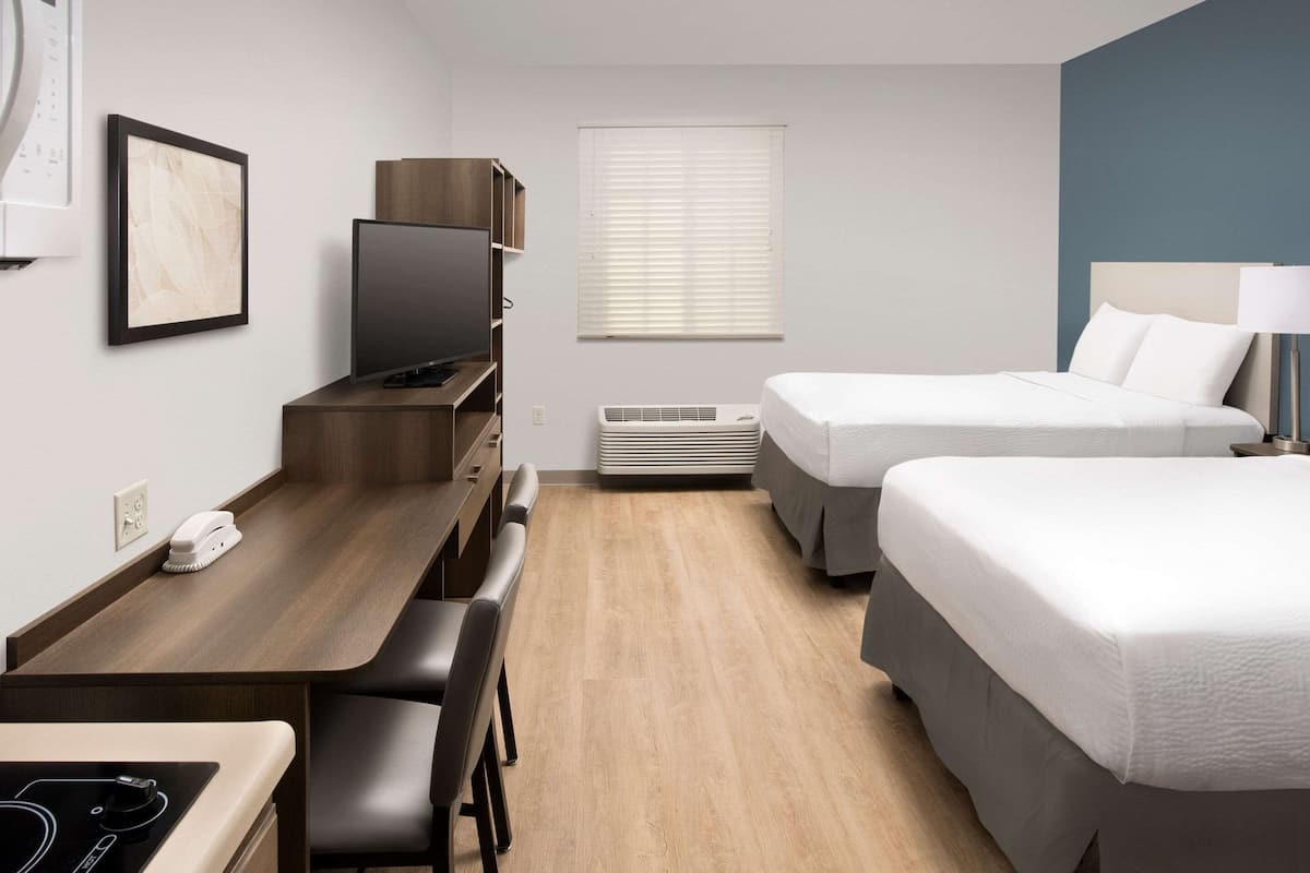Ein Hotelzimmer mit einem Holztisch, einem Fernseher, zwei Betten, einem Fenster mit Jalousien und einer Wandklimaanlage.