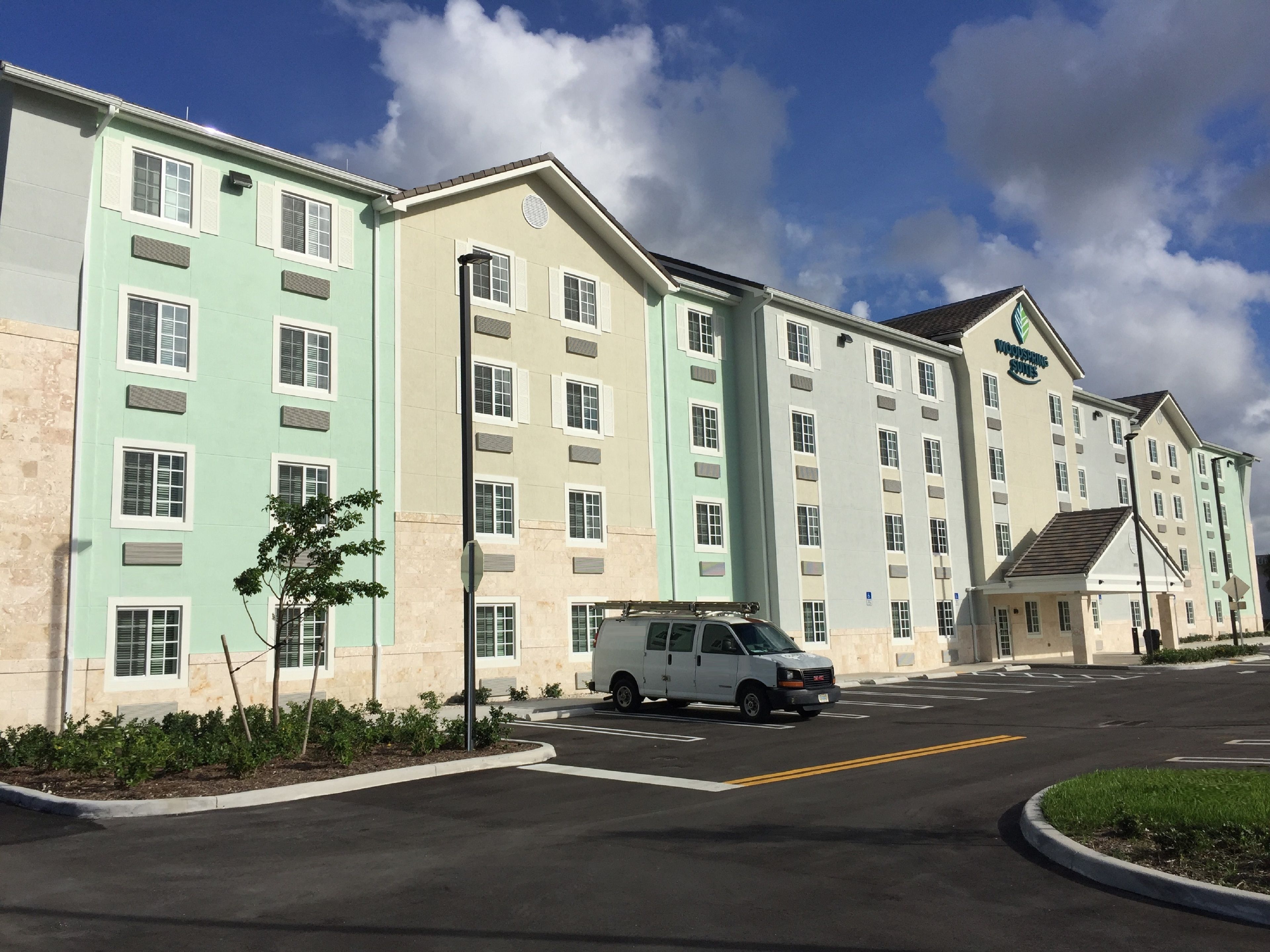 A multi-story hotel with a light green facade, a parking lot, and a white van parked in front.