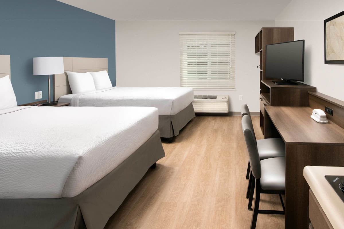 Ein Hotelzimmer mit zwei Betten, einem Schreibtisch mit Stuhl, einem Fernseher und einem Fenster mit Jalousien.