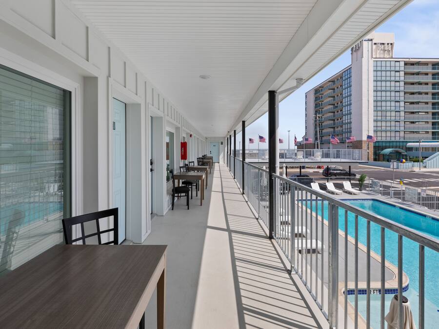 A balcony with a pool, tables, and chairs, overlooking a parking area and a building.