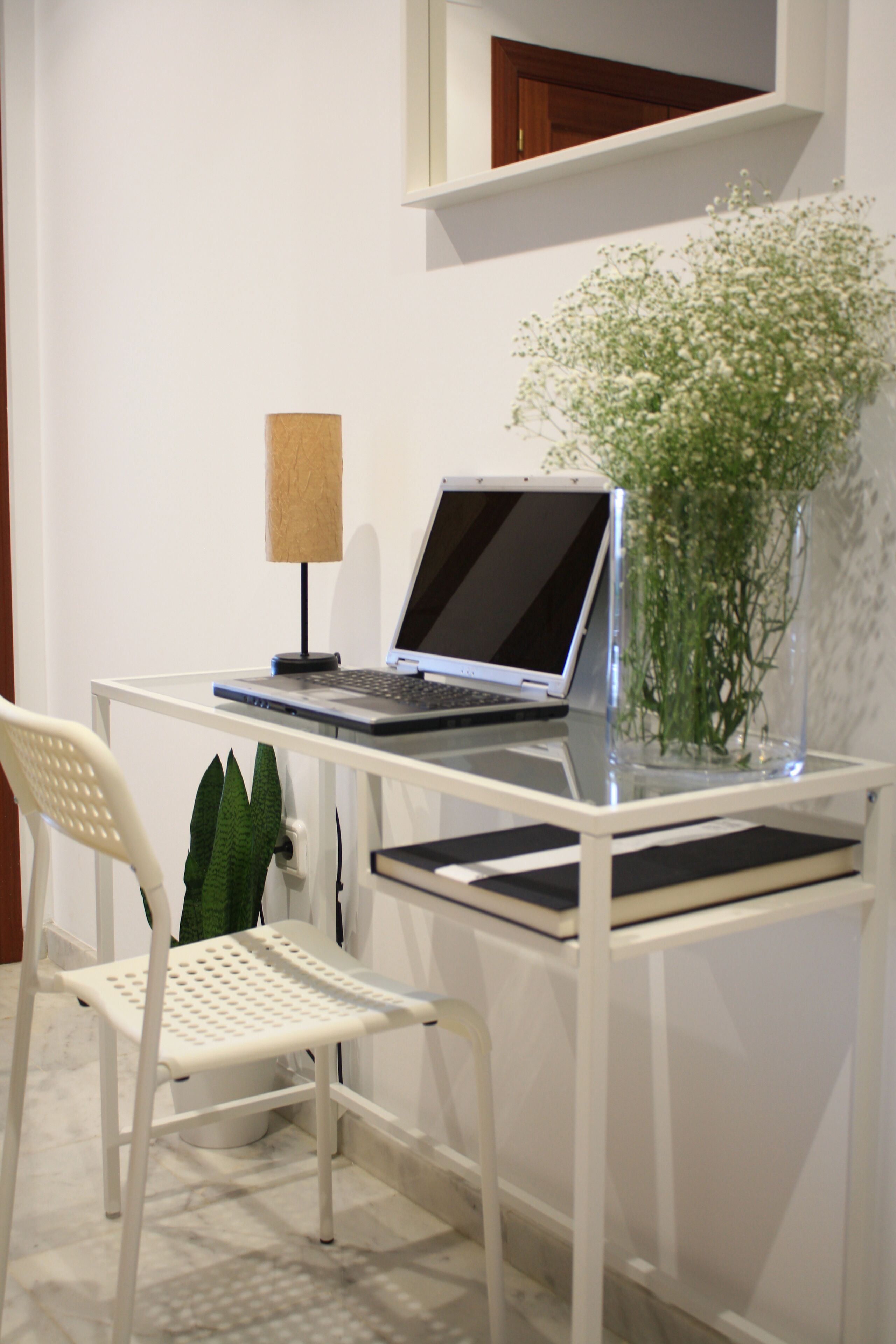 A white desk with a laptop, a lamp, a vase with flowers, and a chair.