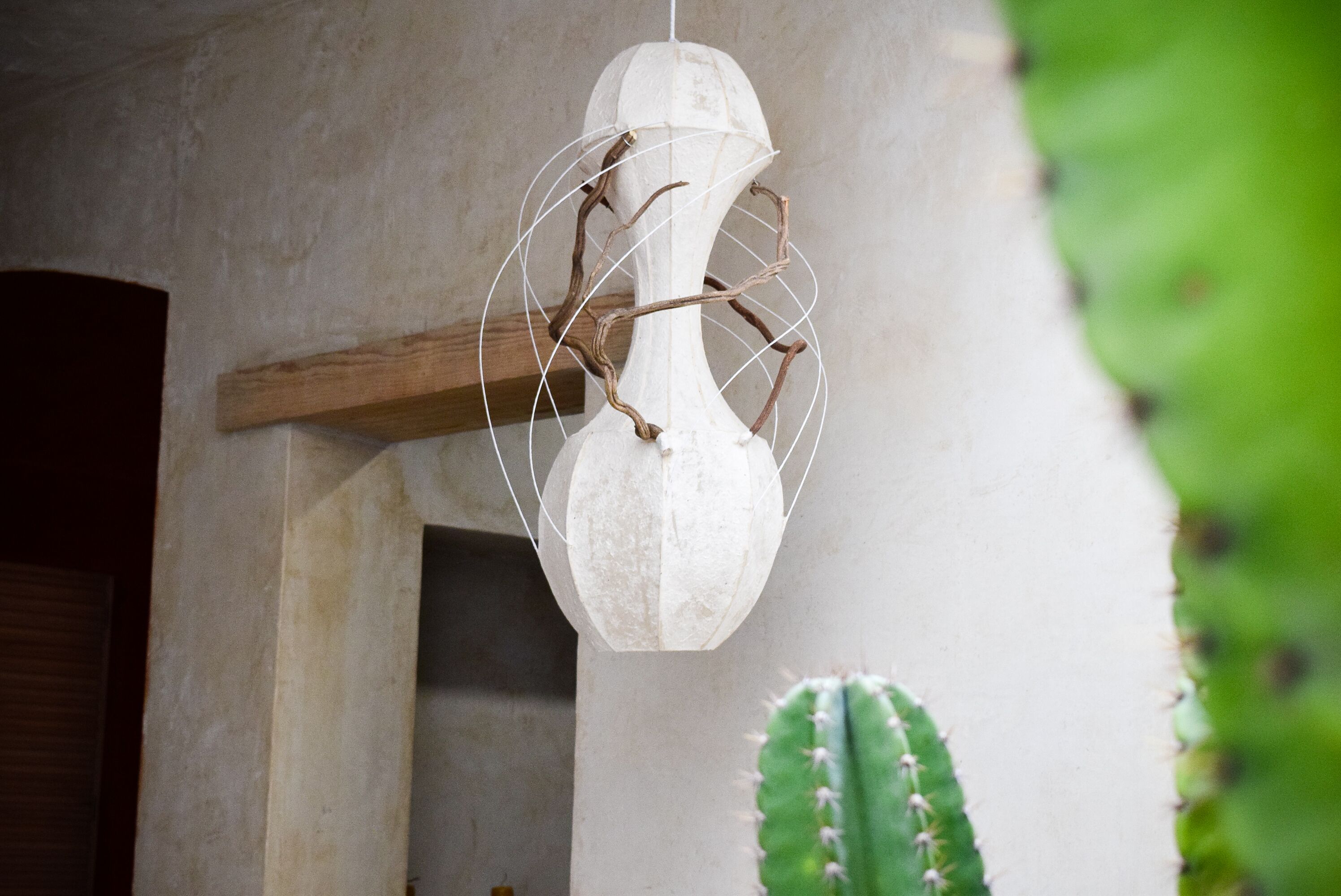 A white, abstract sculpture with branches and a spherical form, mounted on a wall with a wooden beam and a cactus in the background.