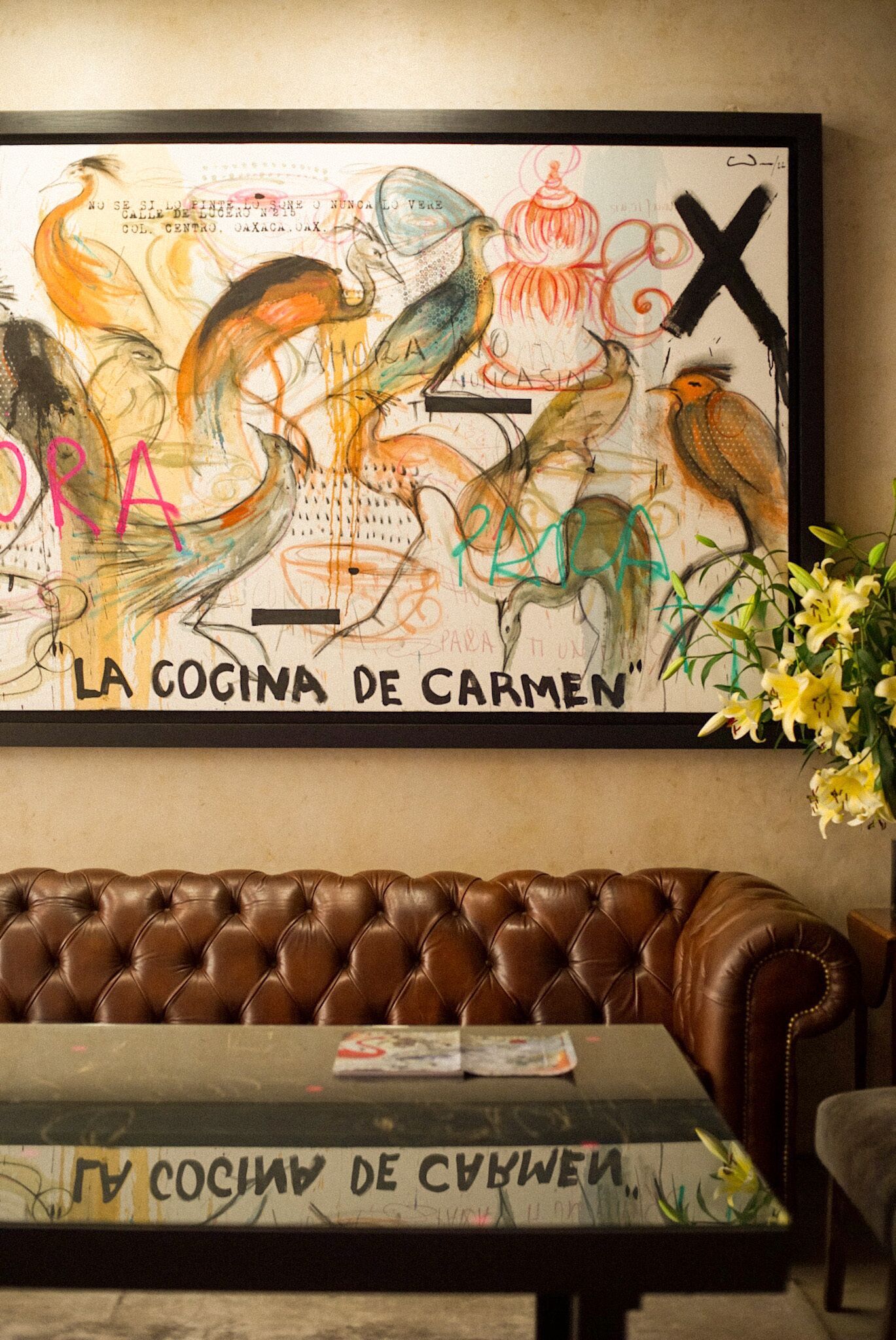 A framed artwork titled "La Cocina de Carmen" above a brown leather couch with a magazine on a table.
