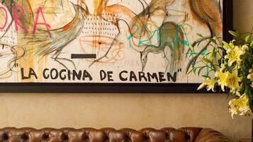 A framed artwork titled "La Cocina de Carmen" above a brown leather couch with a magazine on a table.