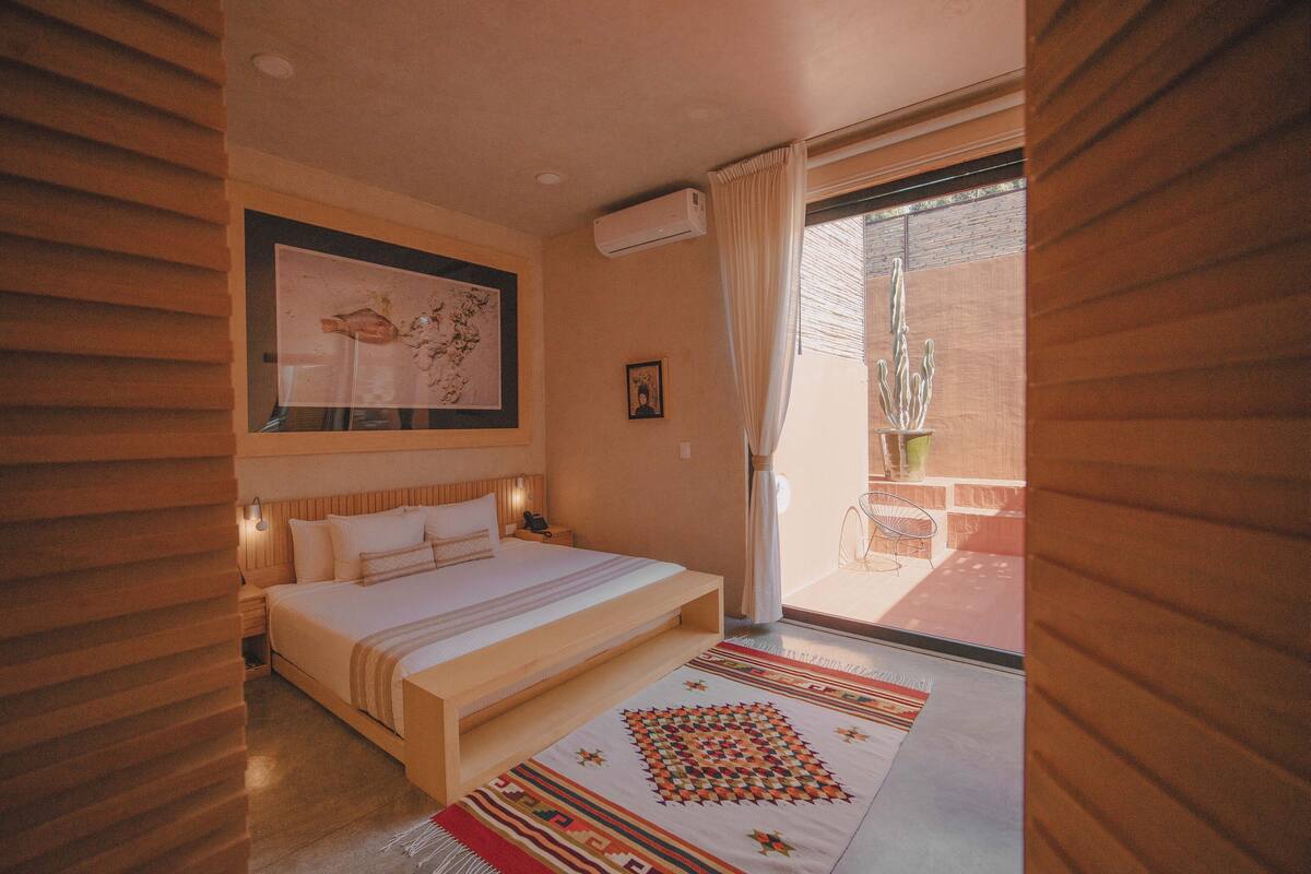 Une chambre à coucher comprenant un lit, une table de chevet, un climatiseur fixé au mur, un tableau encadré et une vue sur un espace extérieur agrémenté d’un cactus.
