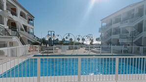 Una piscina con recinzione bianca, un edificio illuminato dal sole con balconi e cielo sereno.