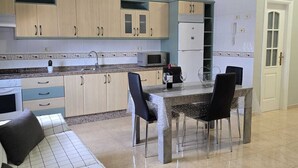 A modern kitchen with light wood cabinets, a microwave, and a refrigerator. There is a dining table with chairs and a sofa with a patterned cover.