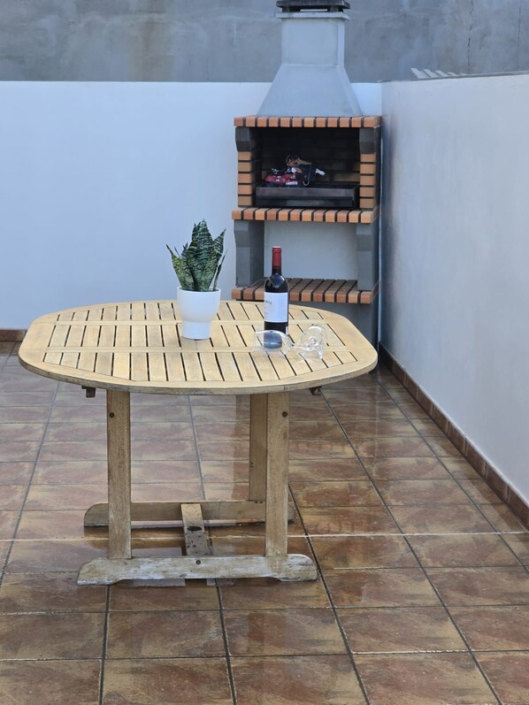 A round wooden table with a potted plant and a bottle of wine on it, placed on a tiled patio with a brick barbecue grill in the background.