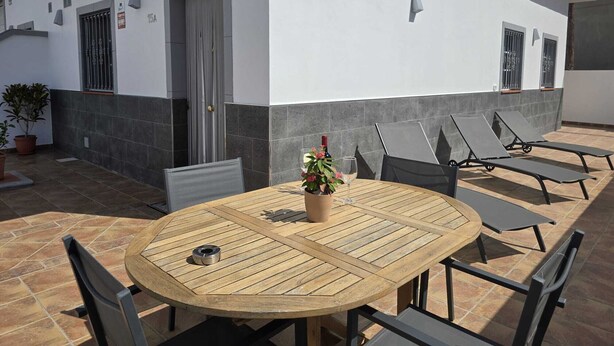 A patio with a round wooden table, two chairs, a potted plant, and a bottle of wine.