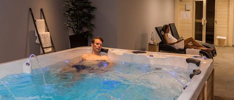 Ein Mann in einer Whirlpool-Badewanne, ein Handtuch liegt daneben, eine Pflanze und ein Stuhl, im Hintergrund liest eine Person.