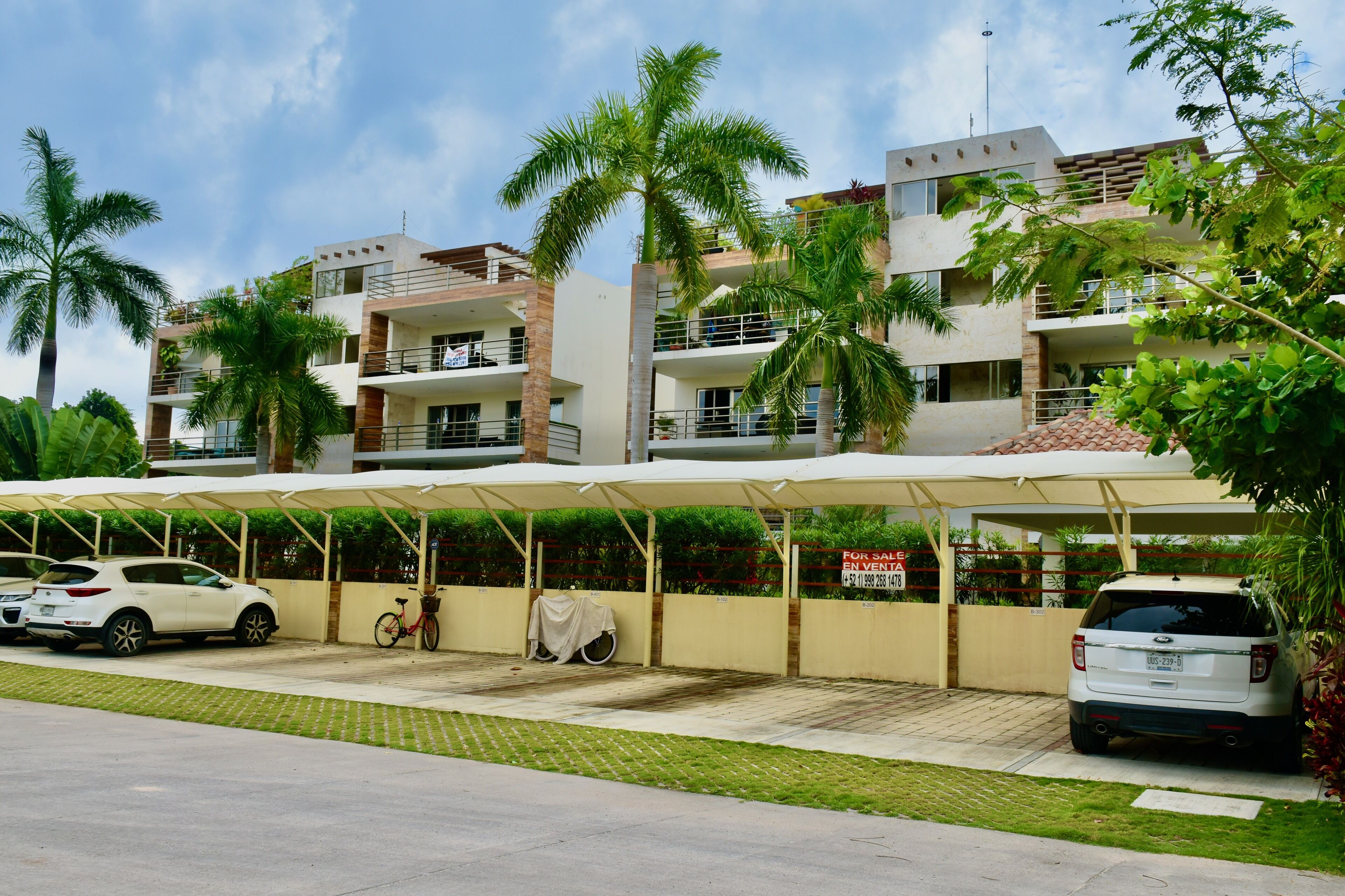A multi-story residential building with balconies, a covered parking area, and palm trees.