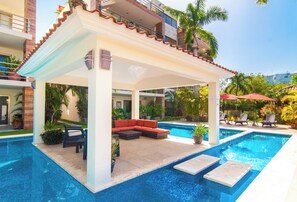 A covered outdoor pool area with a red sofa and lounge chairs, surrounded by potted plants and a clear blue pool.
