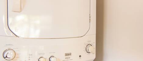 A front-loading white washing machine with control knobs and a digital display, installed in a corner of a room with a window above it.