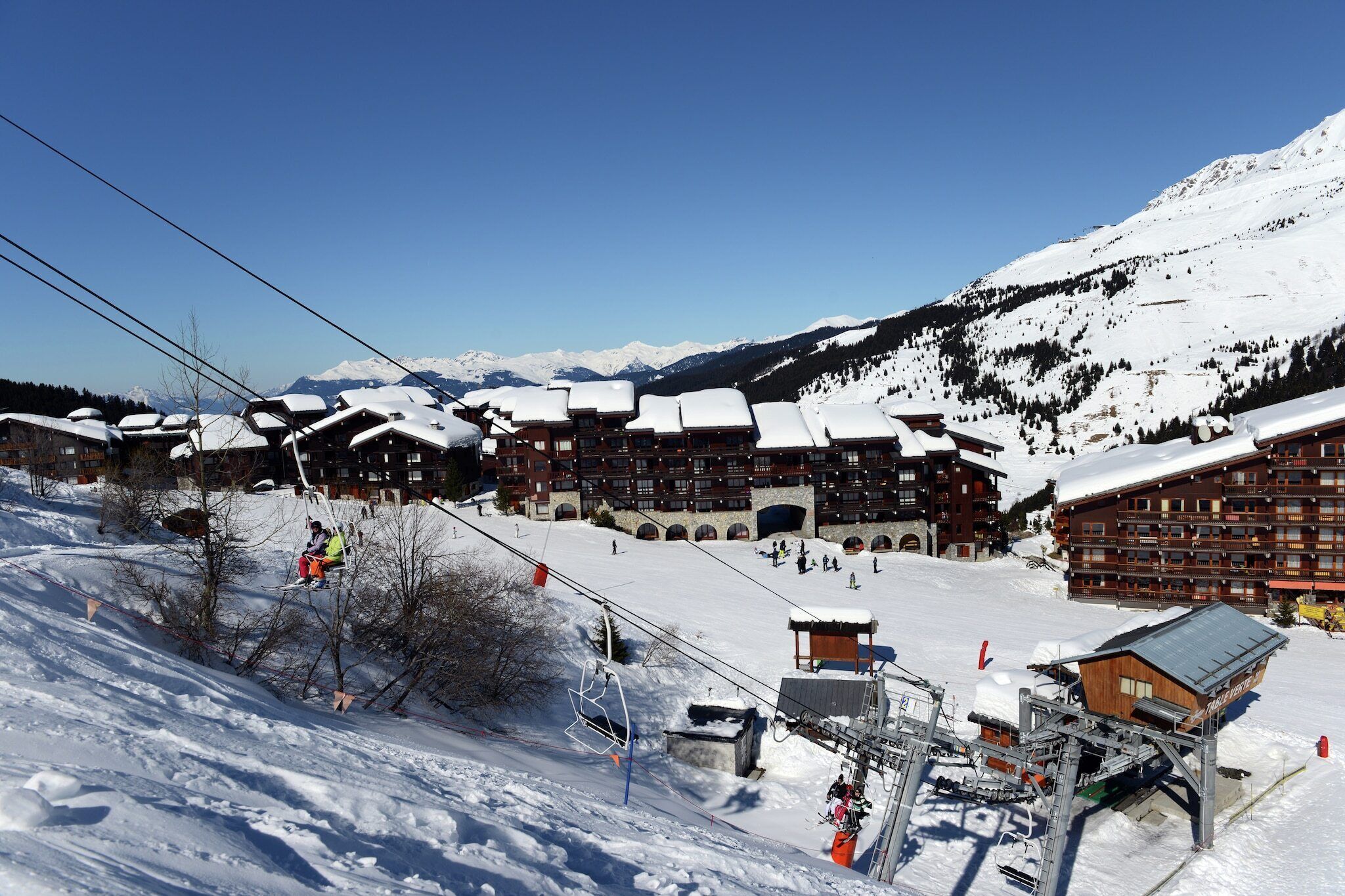 Una estació d'esquí a la muntanya plena de neu, amb diversos edificis de fusta, telesquís i un cel serè.