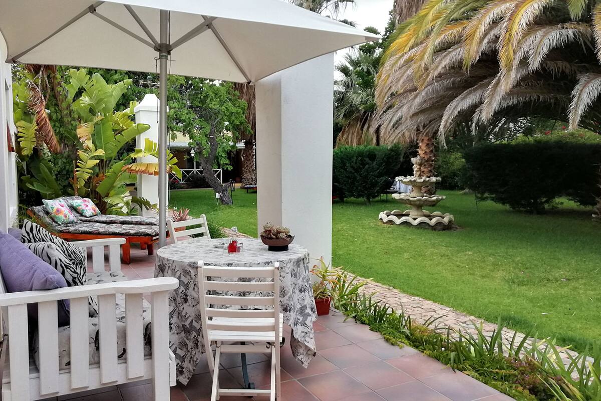 An outdoor patio with a table and chairs, surrounded by lush greenery and a small fountain.