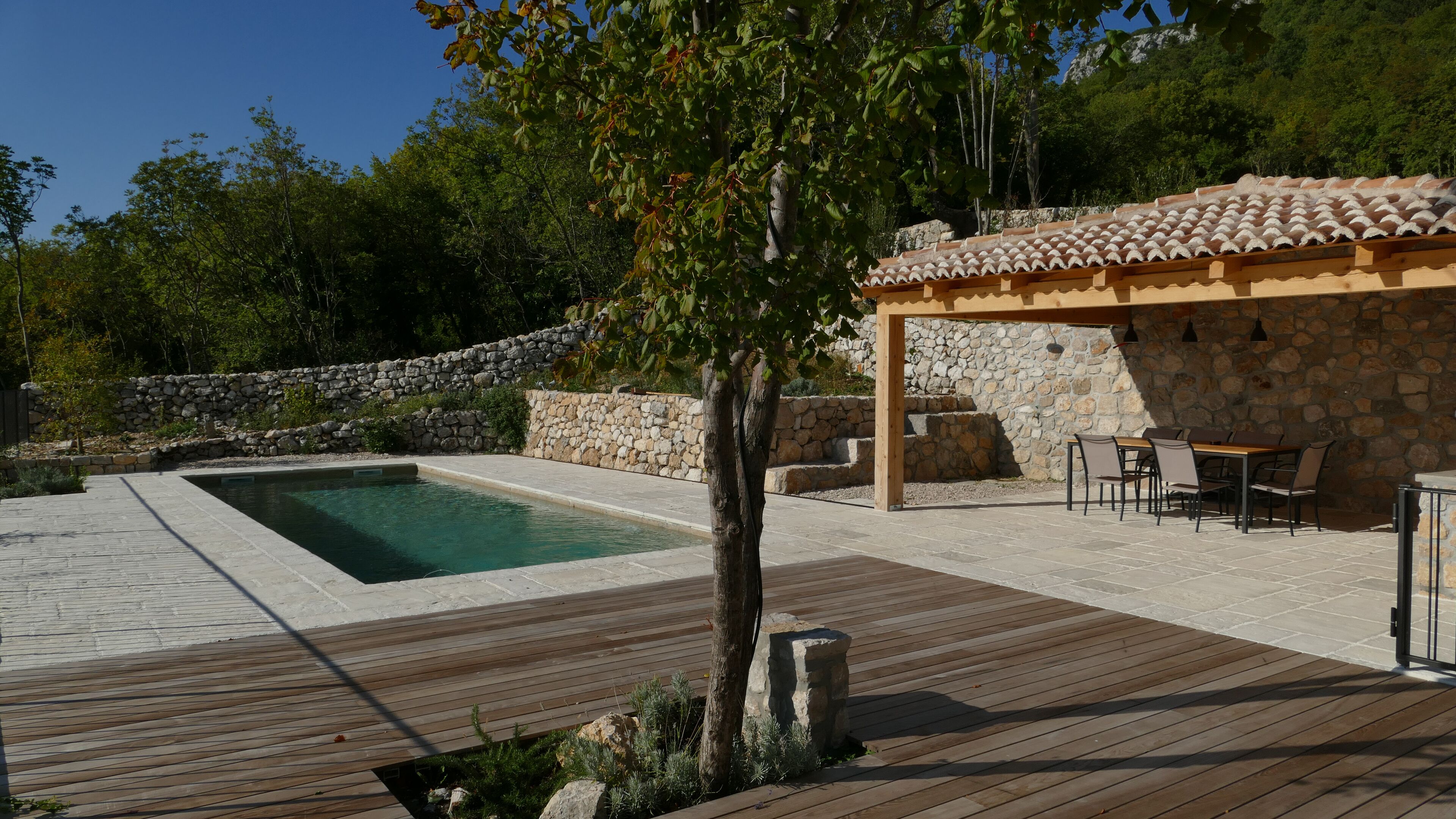 Un espace piscine doté d’une terrasse en bois, de murs en pierre et d’une pergola avec des sièges d’extérieur.