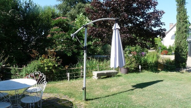 A garden patio with a round table and chairs, surrounded by lush greenery and a white umbrella.