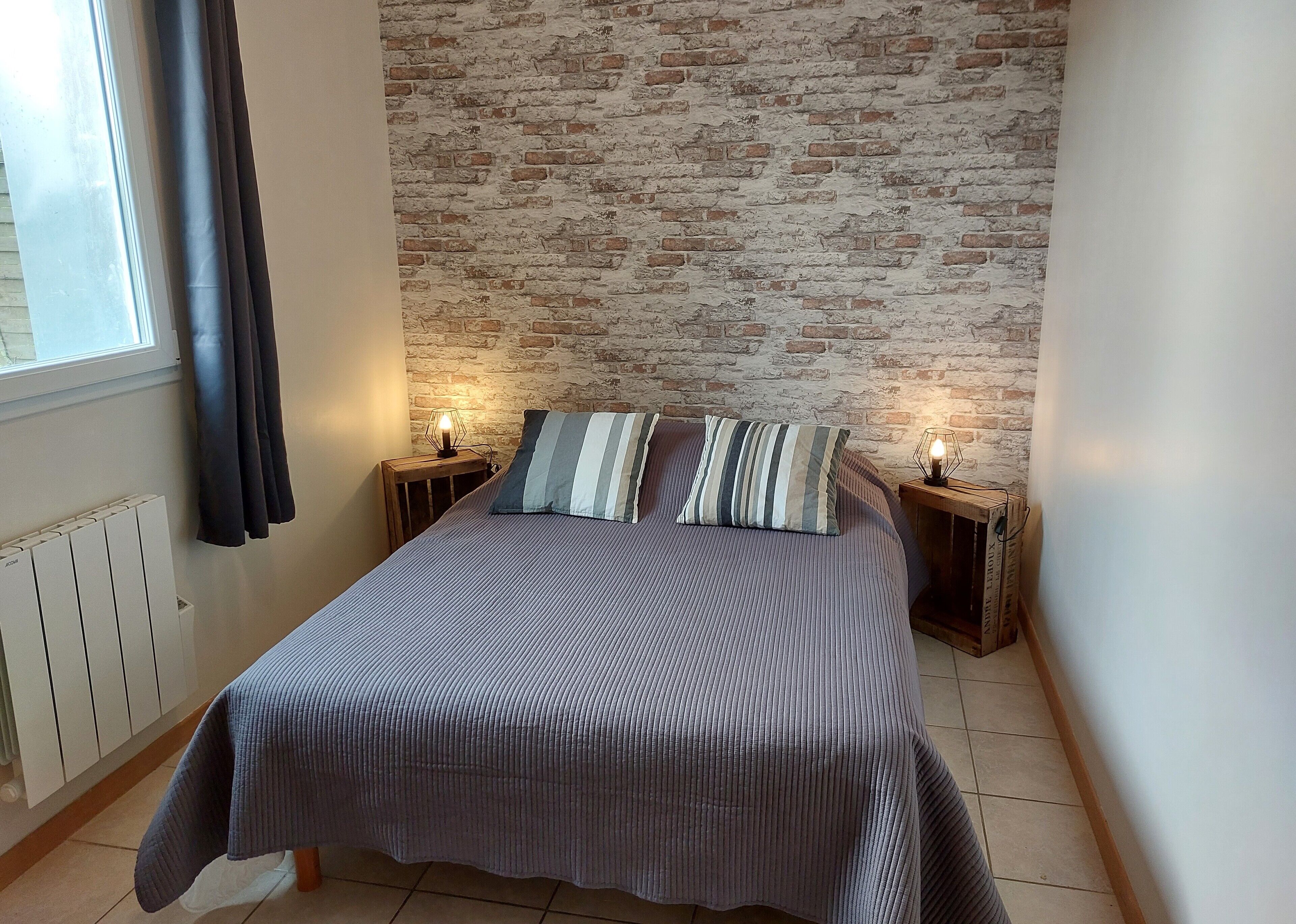 A bedroom with a brick wall, a bed with striped bedding, two bedside lamps, and a window with curtains.