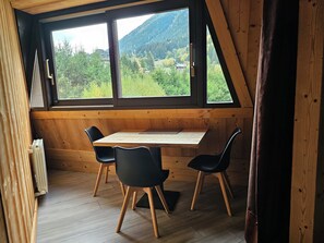 A room with a wooden table and chairs, a window with a view of a forest and mountains, and a radiator on the floor.