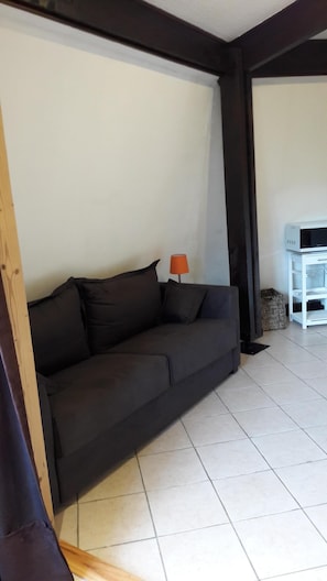 Ein dunkelgraues Sofa vor einer weißen Wand, eine kleine orangefarbene Lampe und ein weißes Schränkchen in einem Raum mit Fliesenboden.