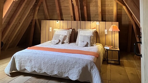 A bedroom with a sloped ceiling, wooden beams, a bed with pillows, a bedside table with a lamp, and a small nightstand. - Le Lapin sur le Toit - Beautifully restored 18th-century barn, private heated swimming pool (Rouffignac-Saint-Cernin-De-Reilhac)