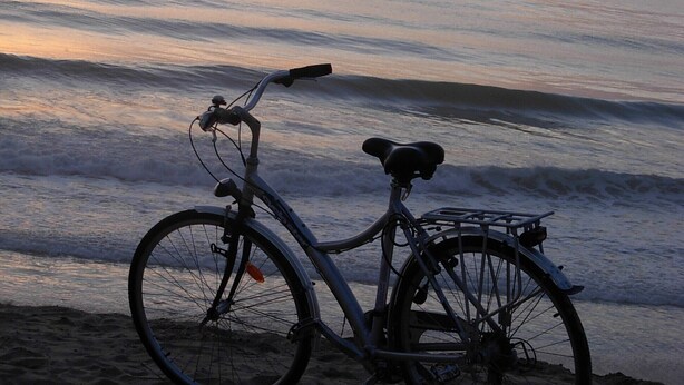 Un vélo garé sur une plage de sable, avec vue sur l’océan et un coucher de soleil en arrière-plan.