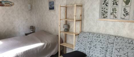 A bedroom with a bed, a sofa, a wooden shelf, a lamp, and a botanical herbarium on the wall.