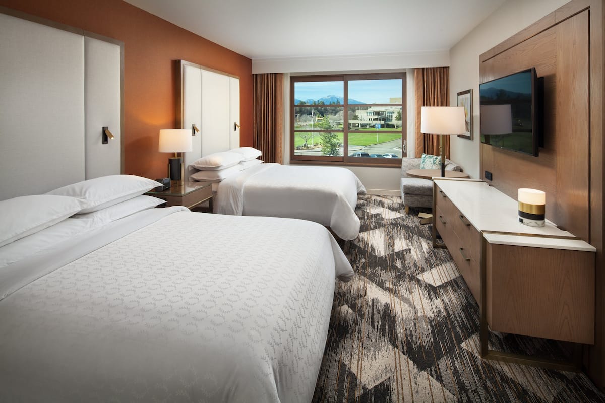 A hotel room with two beds, a desk, a TV, and a large window with a view of a building and mountains.