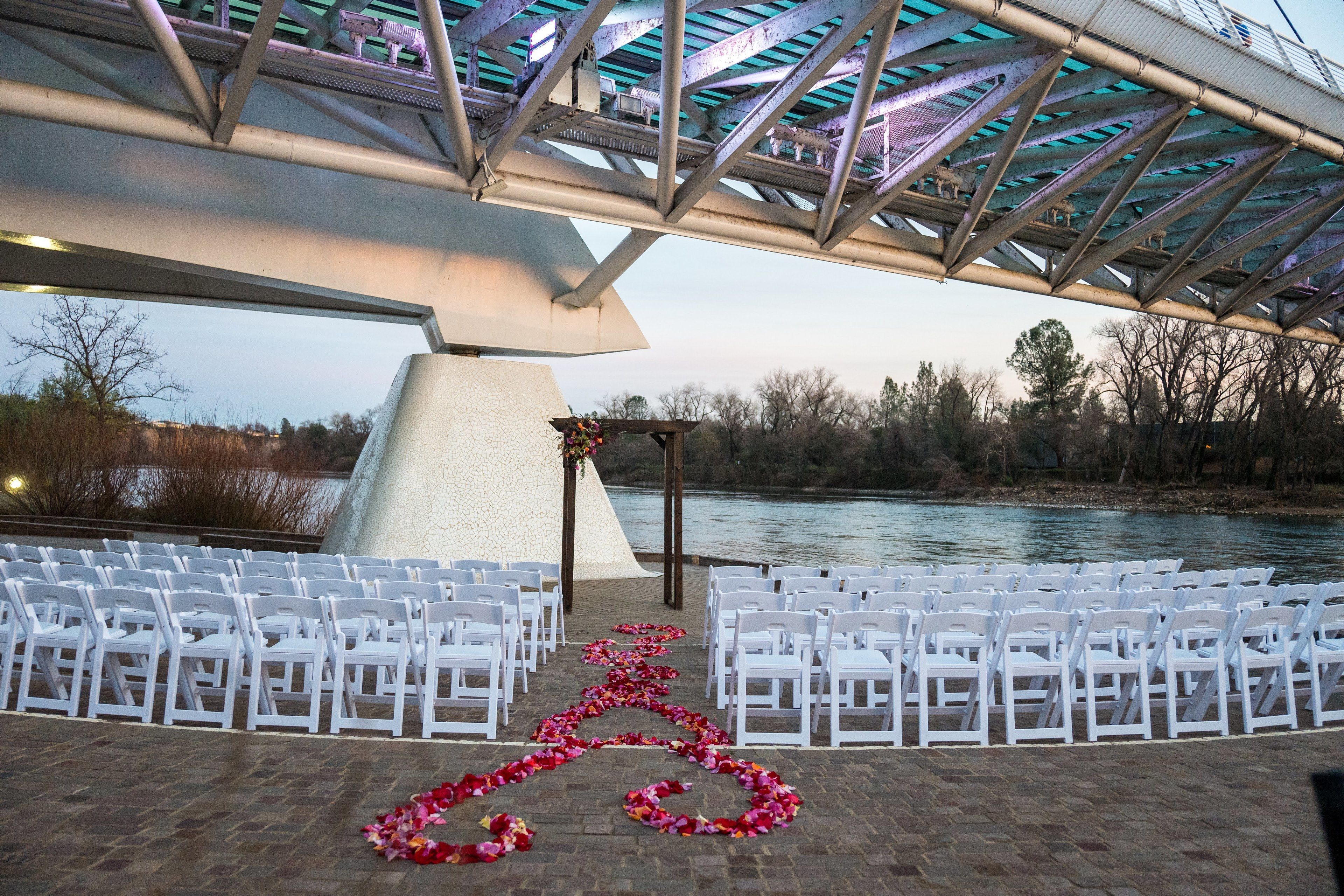 An outdoor wedding ceremony setup with white chairs arranged in rows, a floral arch, and a view of a river.
