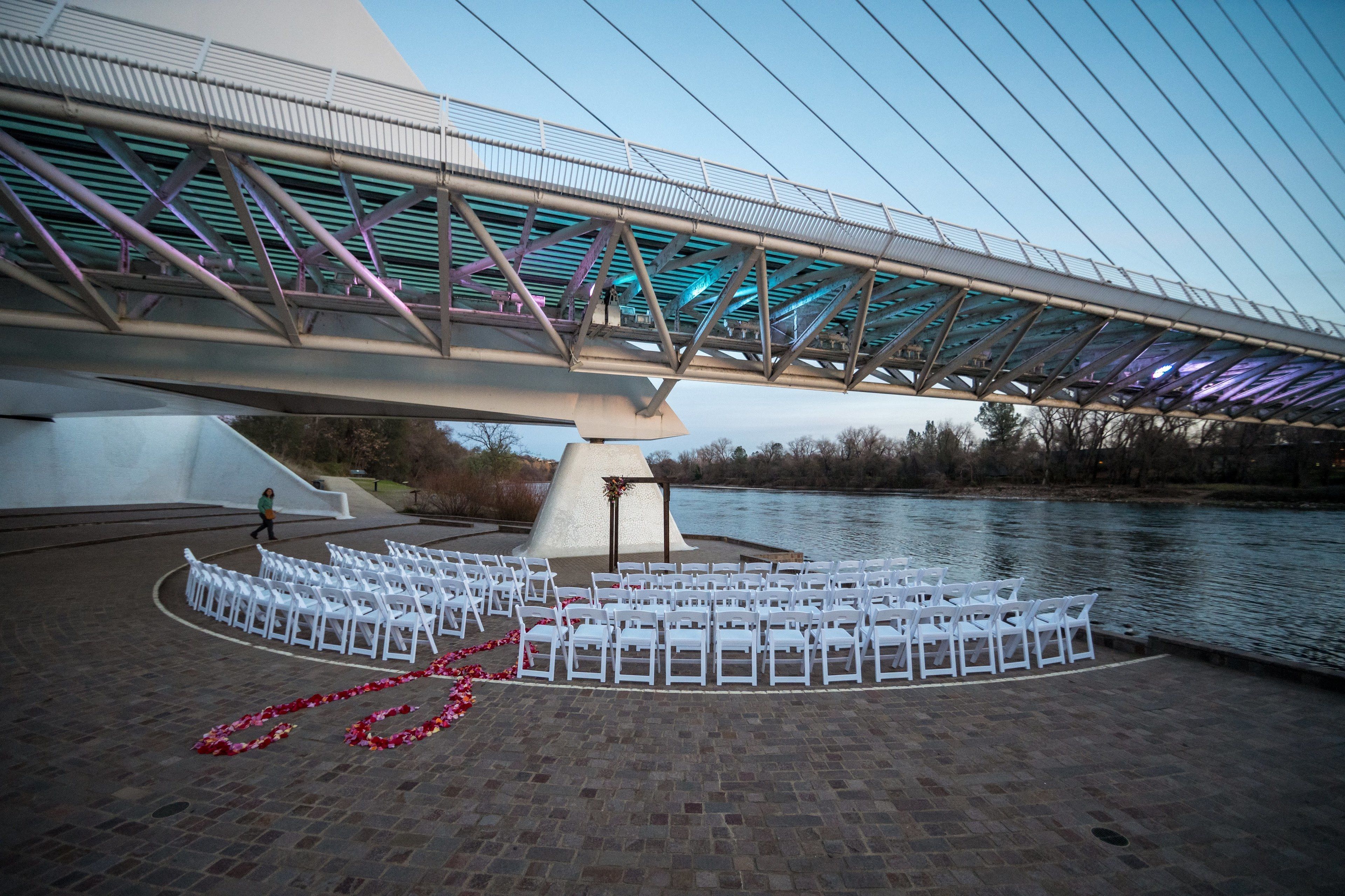 A riverside wedding setup with white chairs arranged in a semi-circle, a bridge overhead, and a decorative floral arch.