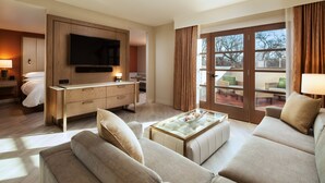 A modern hotel room with a large flat-screen TV, a comfortable sofa, a dining table, and a balcony with seating.