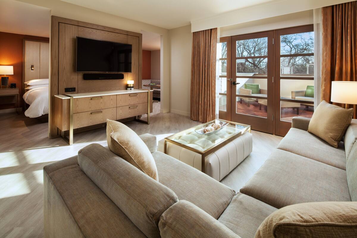 A modern hotel room with a large flat-screen TV, a comfortable sofa, a dining table, and a balcony with seating.