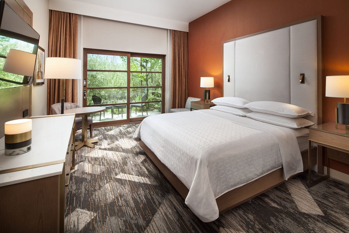 A hotel room with a large bed, a desk, and a chair. There is a window with curtains, a lamp on the desk, and a lamp on the nightstand.