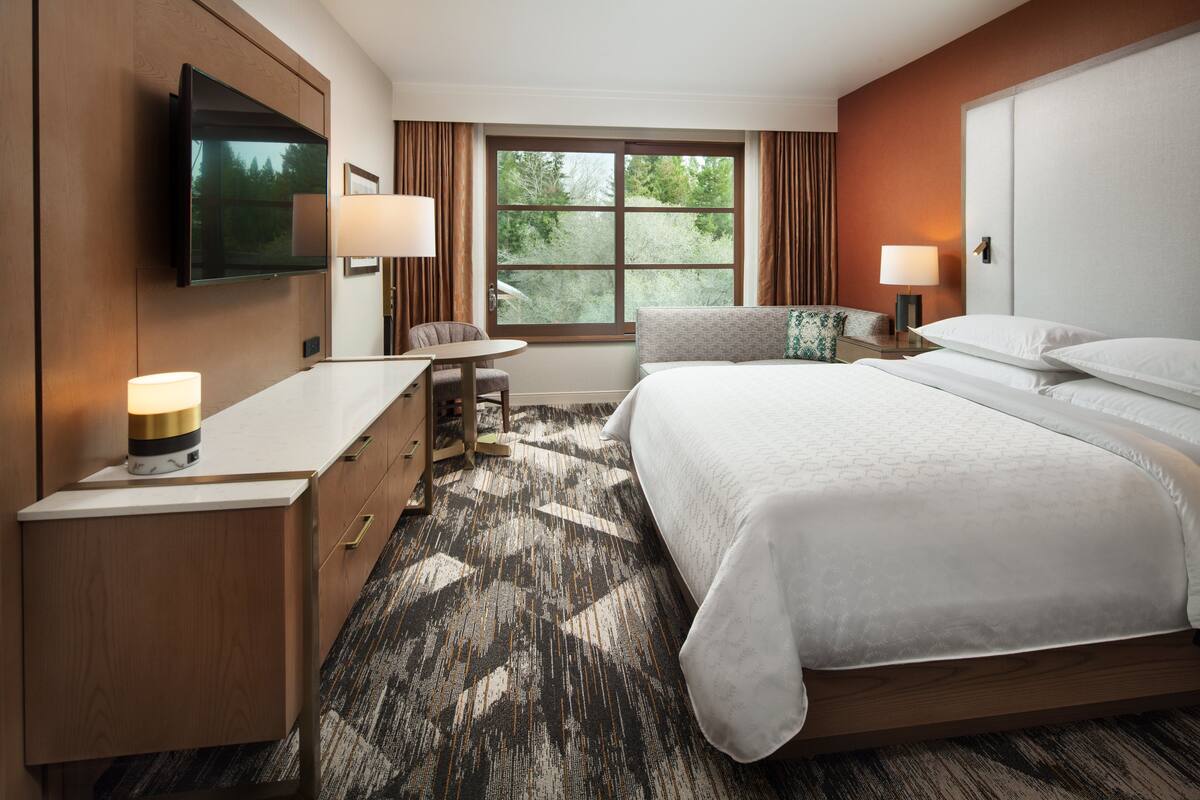 A hotel room with a large bed, a desk, a chair, a TV mounted on the wall, and a window with curtains.