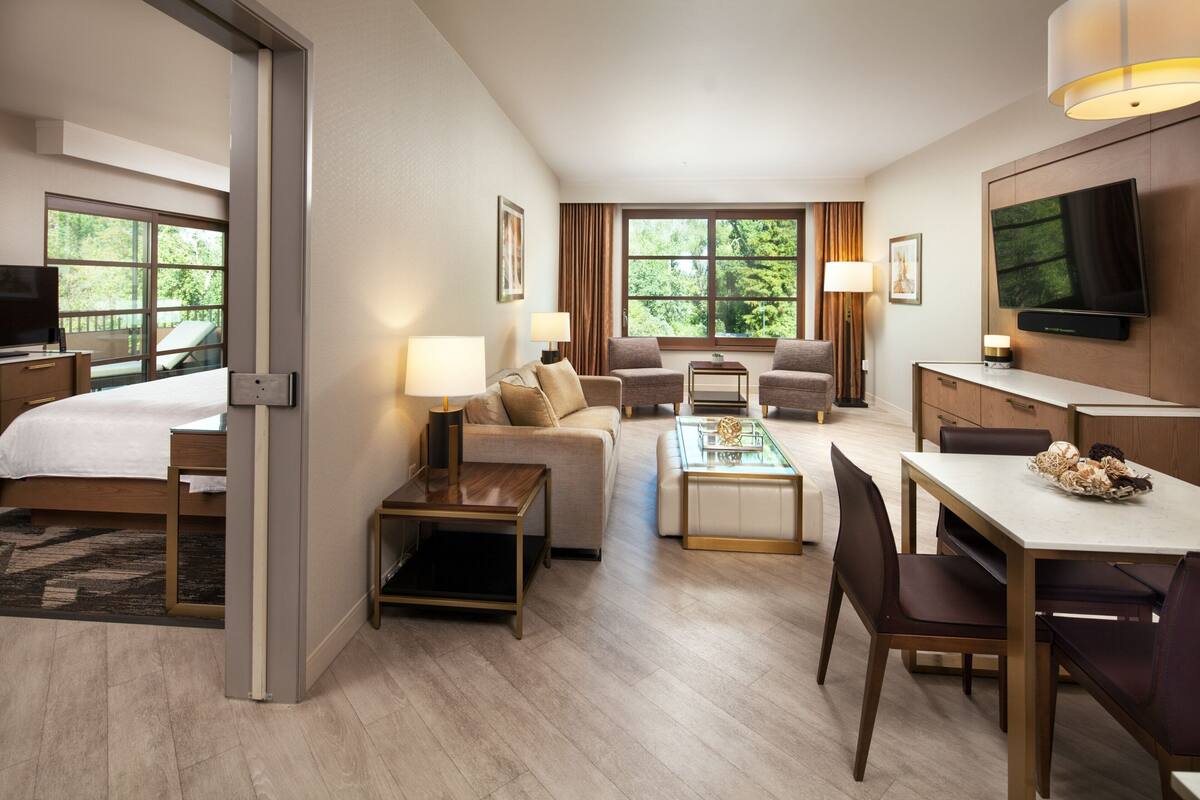 A hotel room with a bed, sofa, armchair, coffee table, dining table, and TV.
