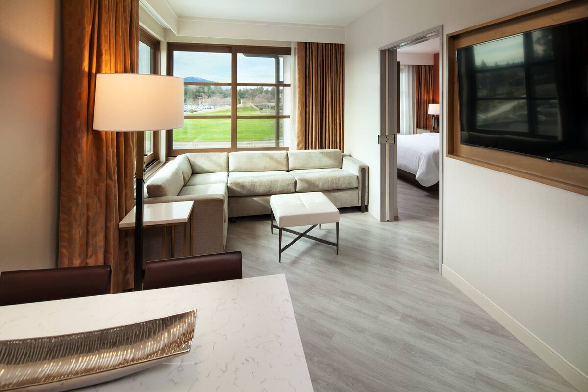A hotel room with a sofa, a dining table, and a view of a green landscape through large windows.