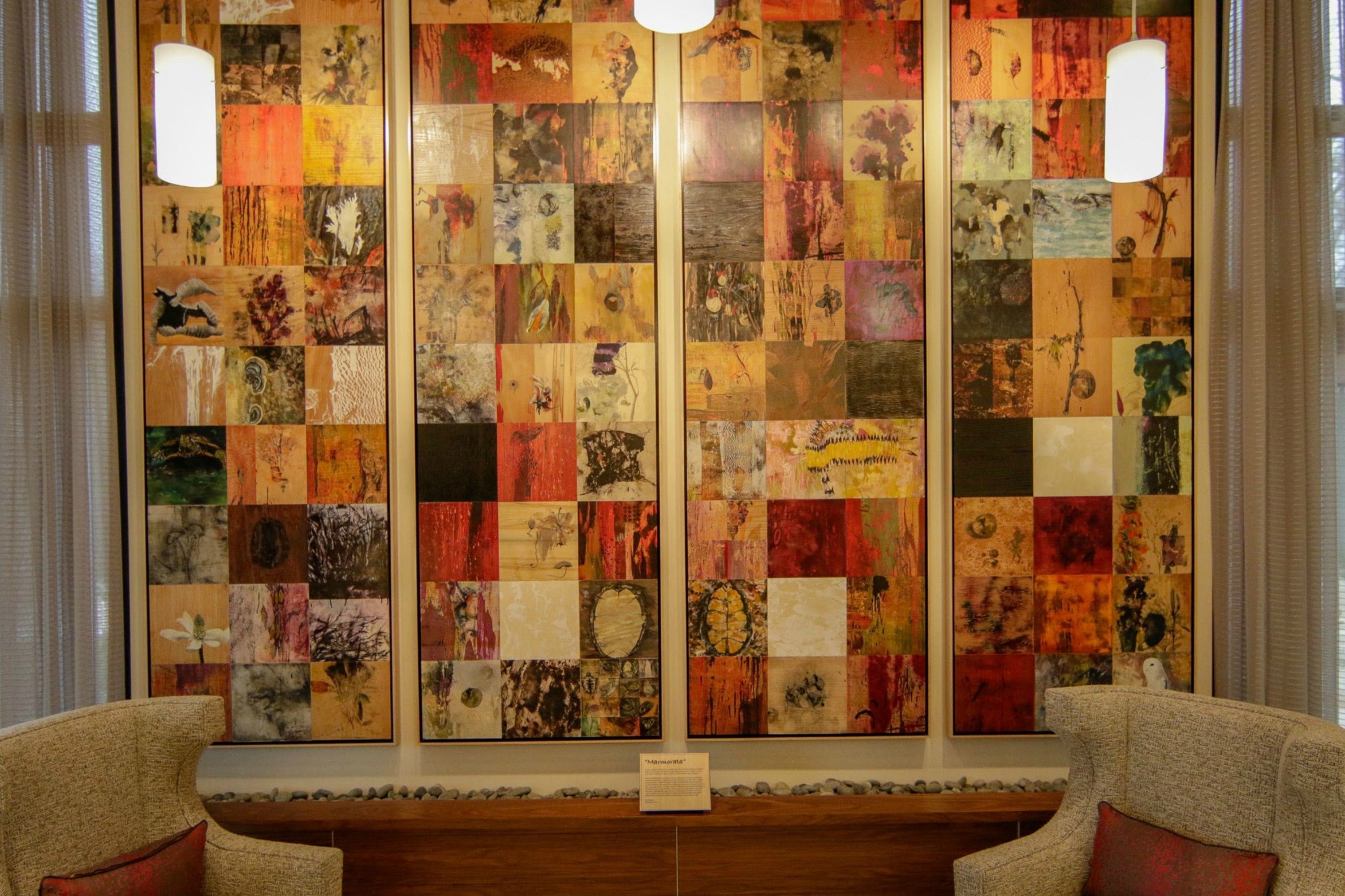 A room with a large, colorful collage artwork on the wall, a wooden bench, and two armchairs.