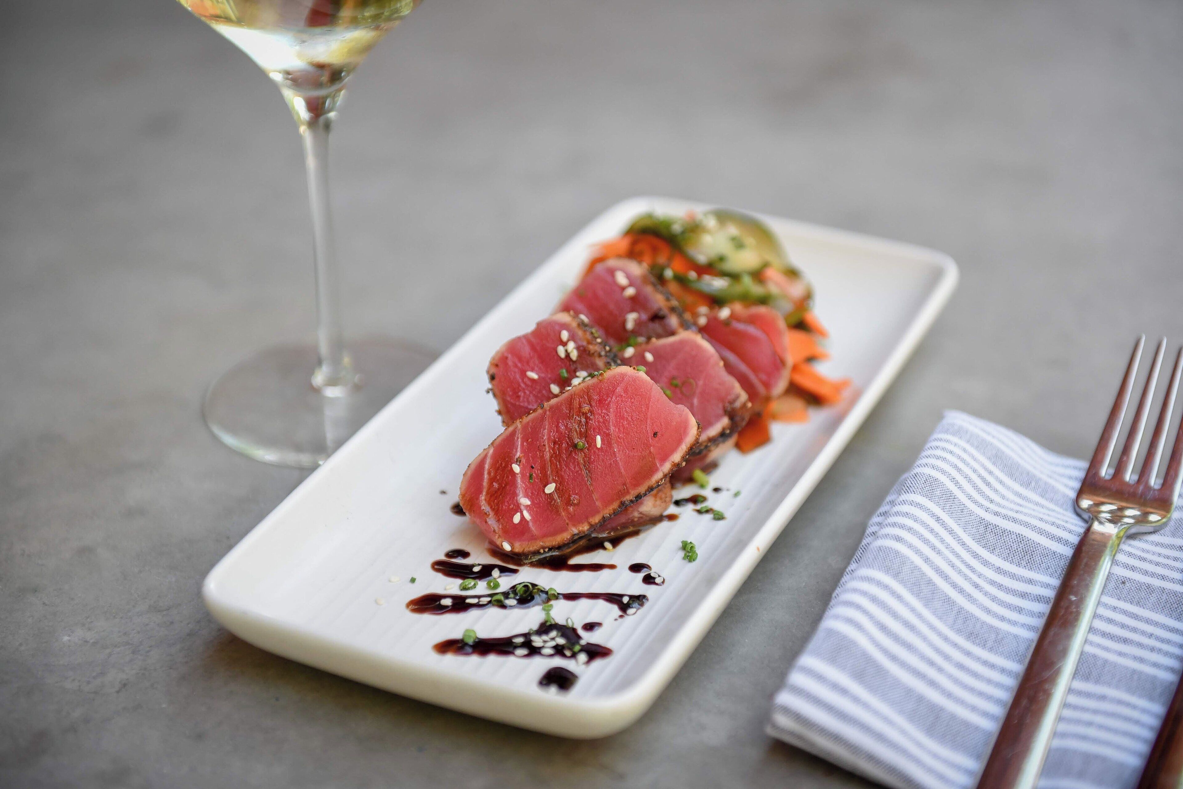 A white rectangular plate with a dish of sliced meat, vegetables, and a drizzle of sauce, accompanied by a glass of white wine and a striped napkin with a fork.