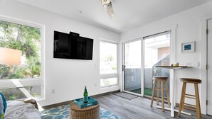 En moderne stue med et stort TV, en skydedør til en balkon og et lille køkkenområde med barstole.