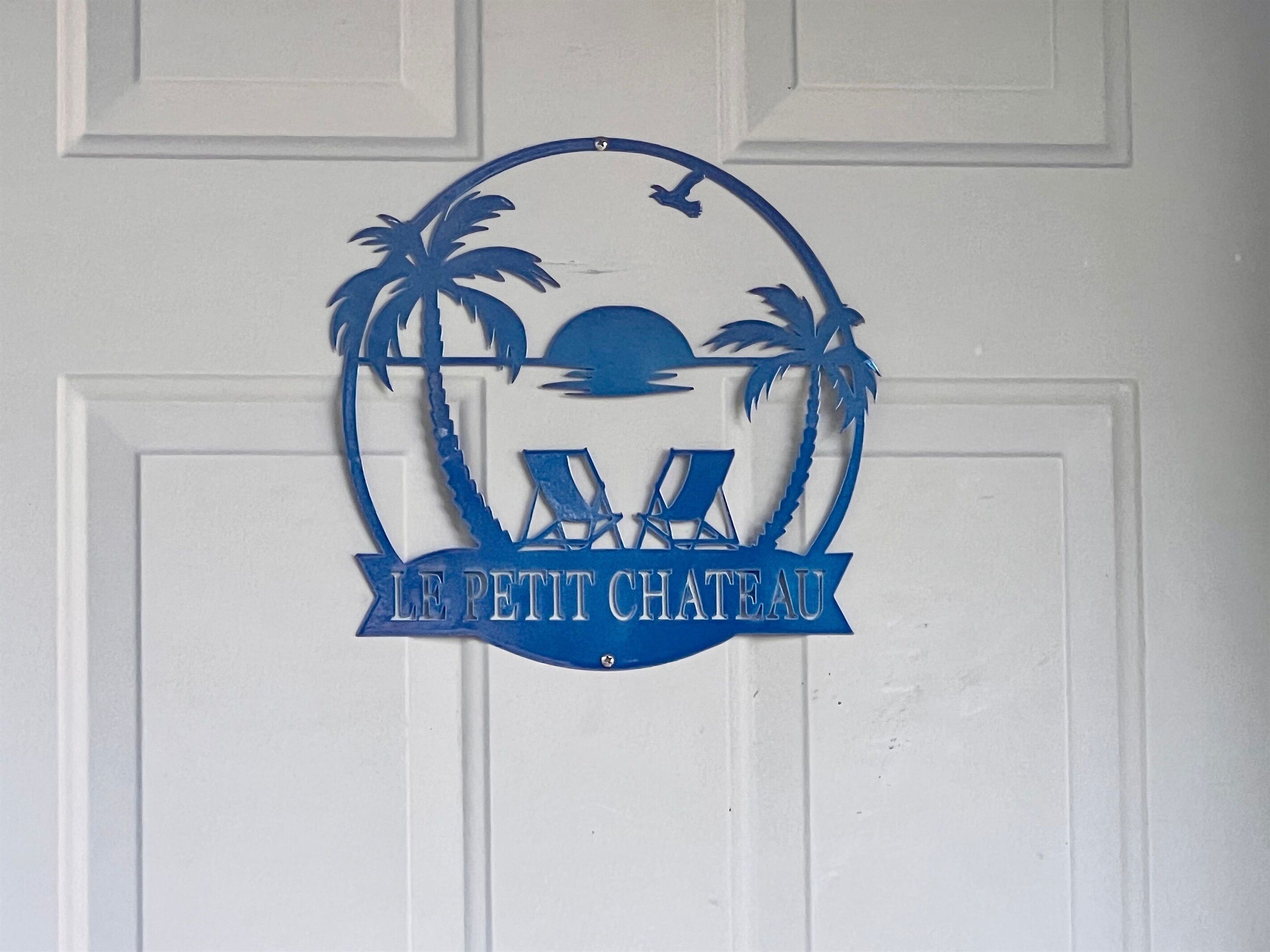 A decorative sign with palm trees, a sun, and a lounge chair, with the text "LE PETIT CHATEAU" on a white door.